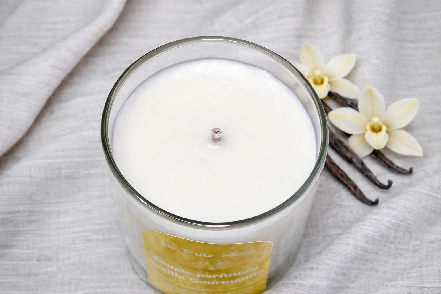 Vue de dessus d'une bougie naturelle Vanille Gourmande avec mèche en coton, posée sur un tissu en lin avec gousses et fleurs