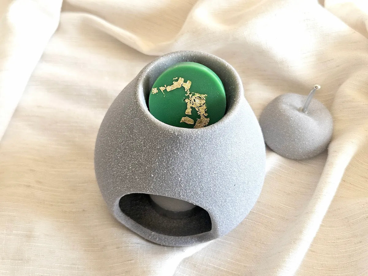 Vue à trois quarts d'un brûleur de parfum en ciment gris clair contenant le palet fondant vert citron vert avec feuille d'or, posé sur une surface de lin.