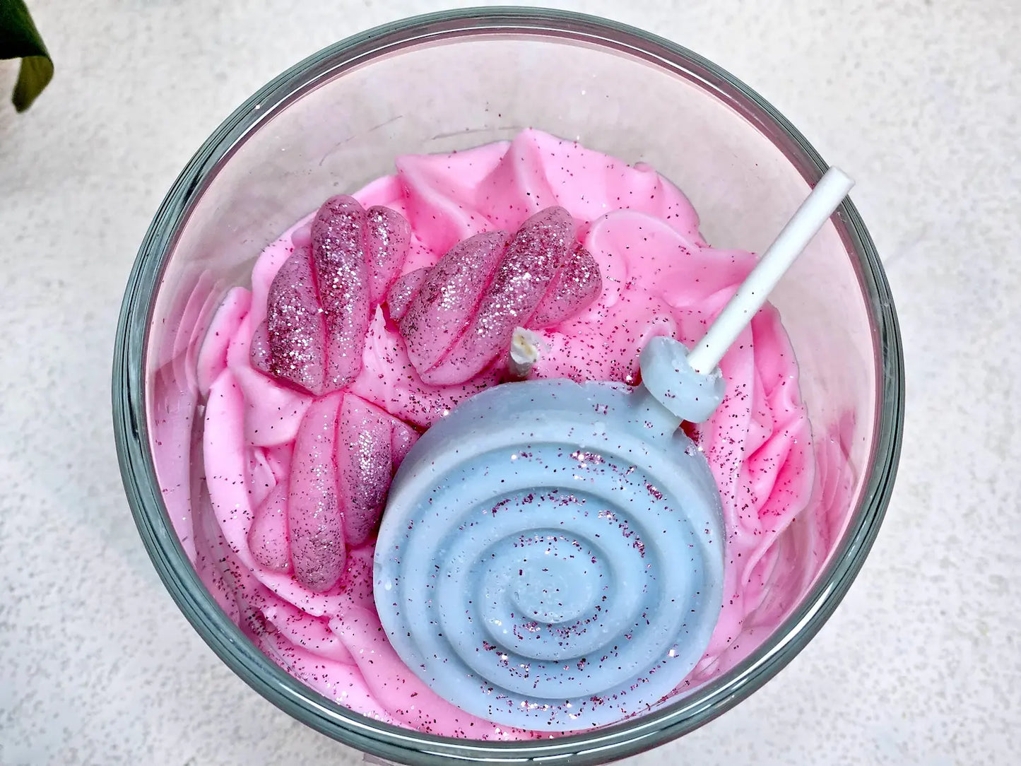 Détail du dessus d'une bougie gourmande avec sucette bleue sculptée en cire et paillettes sur chantilly rose.