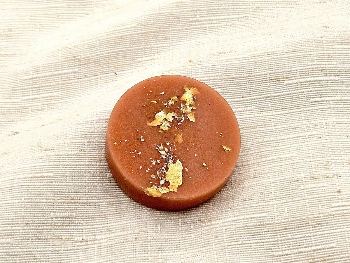Palet fondant parfumé au Lait d'Amande, couleur terre cuite douce avec éclats de feuille d'or, posé sur un tissu de lin clair.