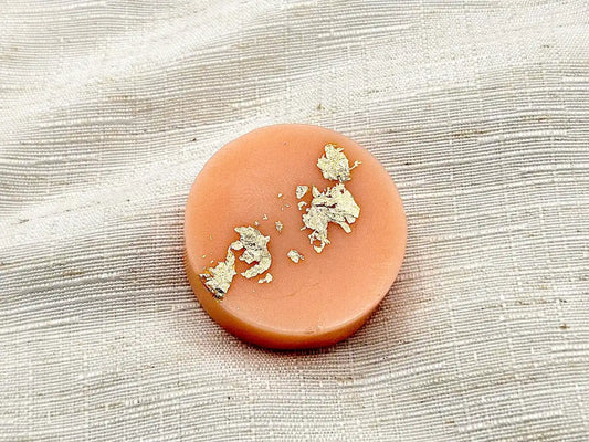 Palet fondant parfumé au Magnolia, de couleur pêche pastel avec éclats de feuille d'or, posé sur un tissu de lin blanc.