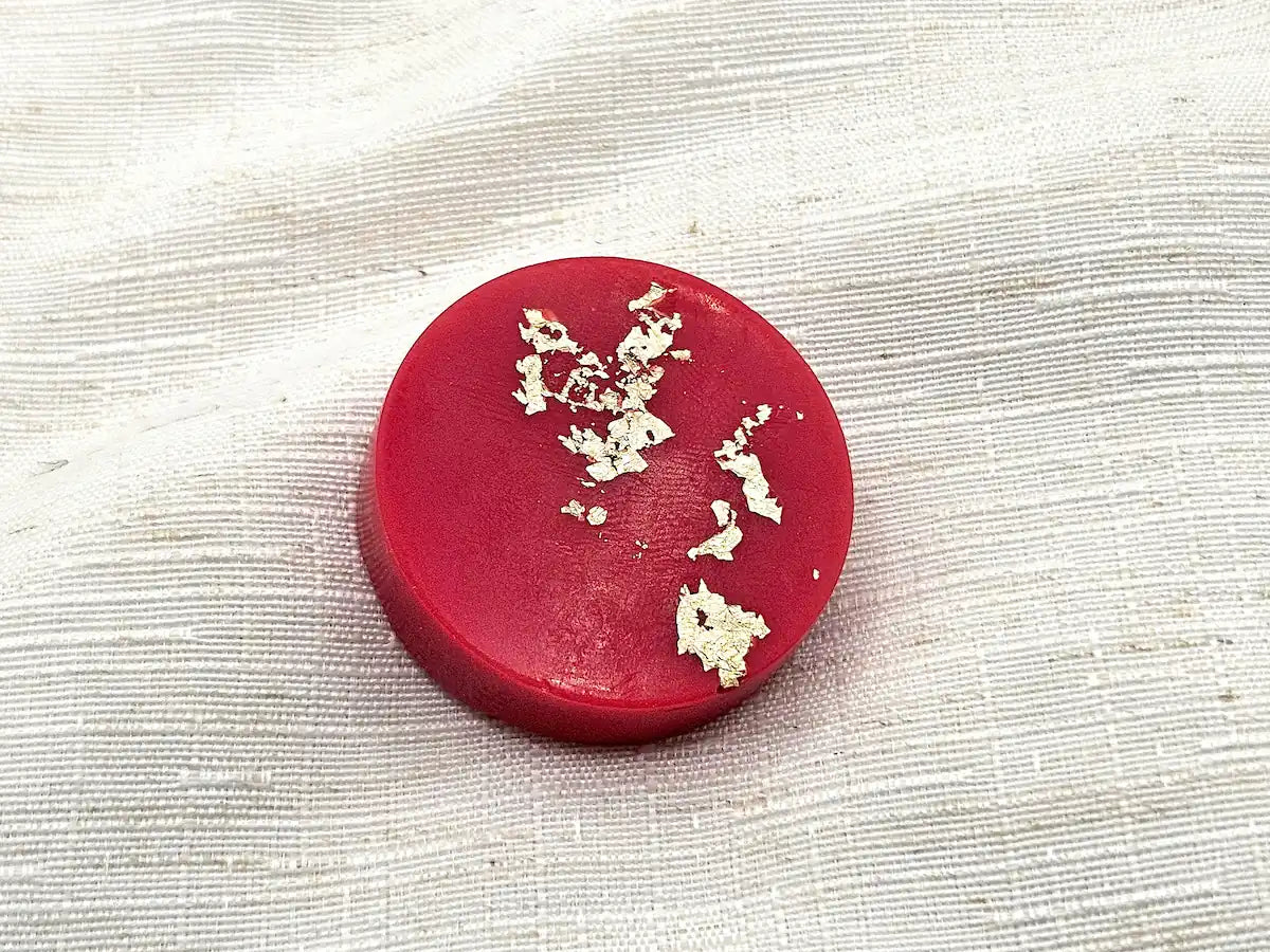 Palet fondant parfumé à la Rhubarbe et Fraise, couleur rouge intense avec éclats de feuille d'or, posé sur un lin texturé.