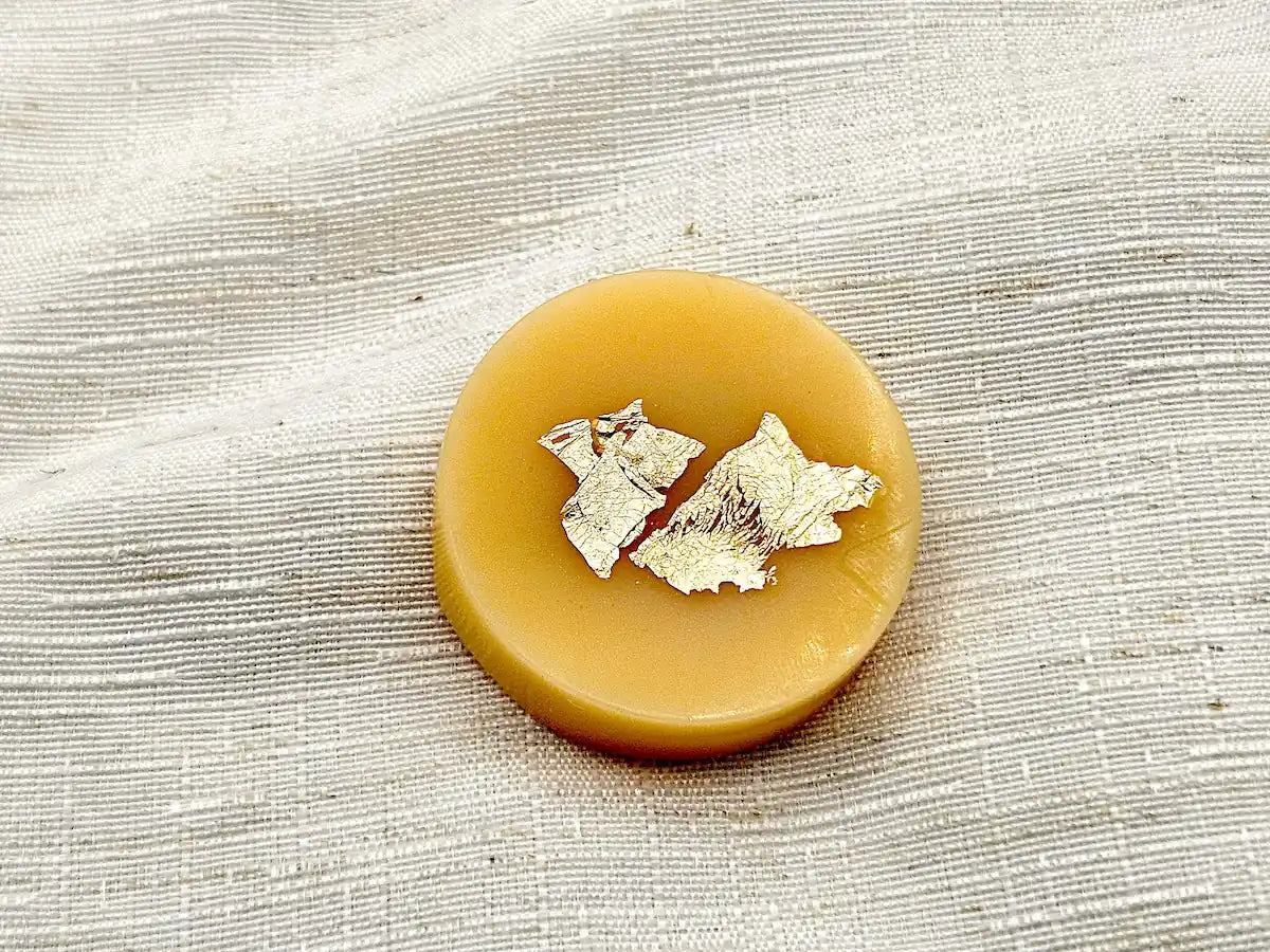 Palet fondant parfumé à la Vanille des Îles, couleur jaune miel avec éclats de feuille d'or, posé sur un tissu de lin blanc.