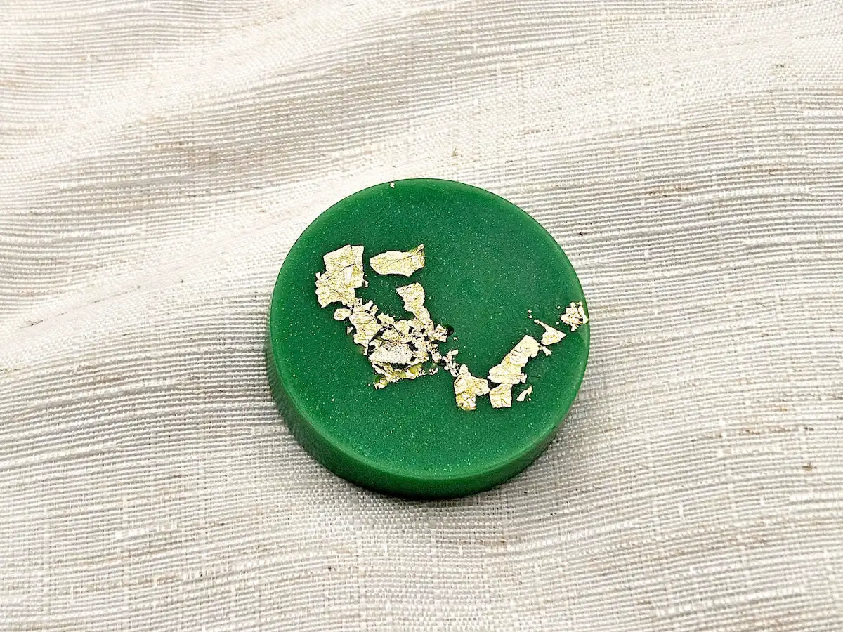 Gros plan détaillé sur un palet fondant parfumé Citron Vert de couleur verte, orné de délicates paillettes de feuille d'or, posé sur un tissu de lin texturé.