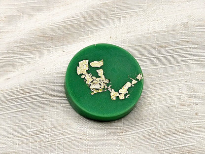 Gros plan détaillé sur un palet fondant parfumé Citron Vert de couleur verte, orné de délicates paillettes de feuille d'or, posé sur un tissu de lin texturé.