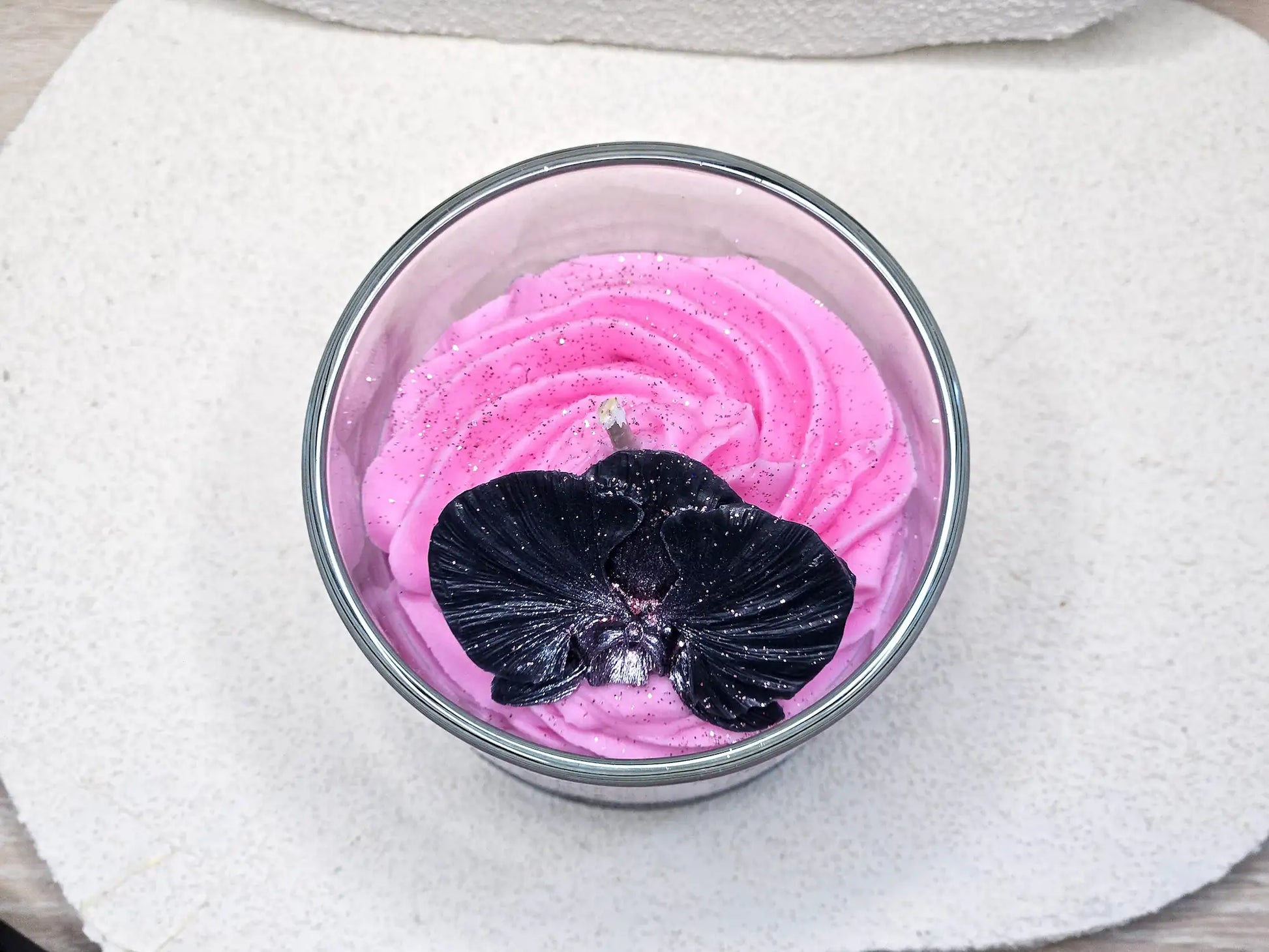 Détail du dessus d'une bougie gourmande avec une fleur d'orchidée noire sculptée en cire sur un lit de chantilly rose.