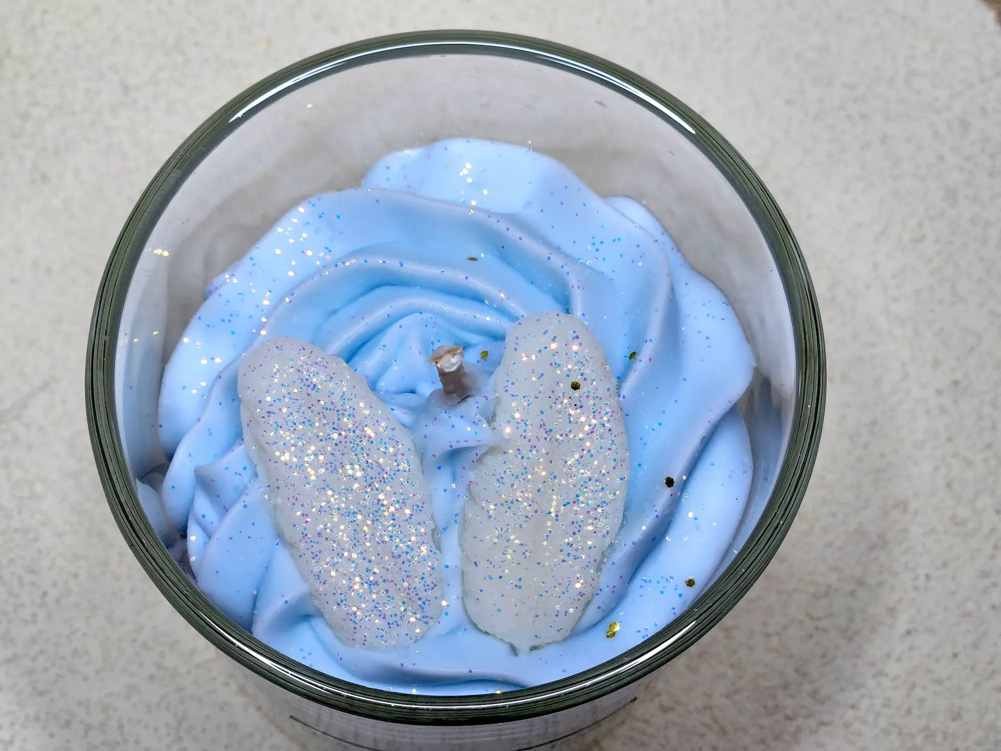 Gros plan sur la cire fouettée bleue et les fleurs décoratives pailletées d'une bougie gourmande parfumée à la fleur de coton.